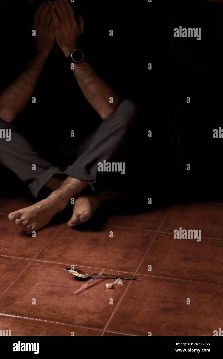 Homme pieds nus non identifié assis sur le sol en semi-obscurité avec une seringue et une cuillère contenant de l'héroïne au premier plan Banque D'Images