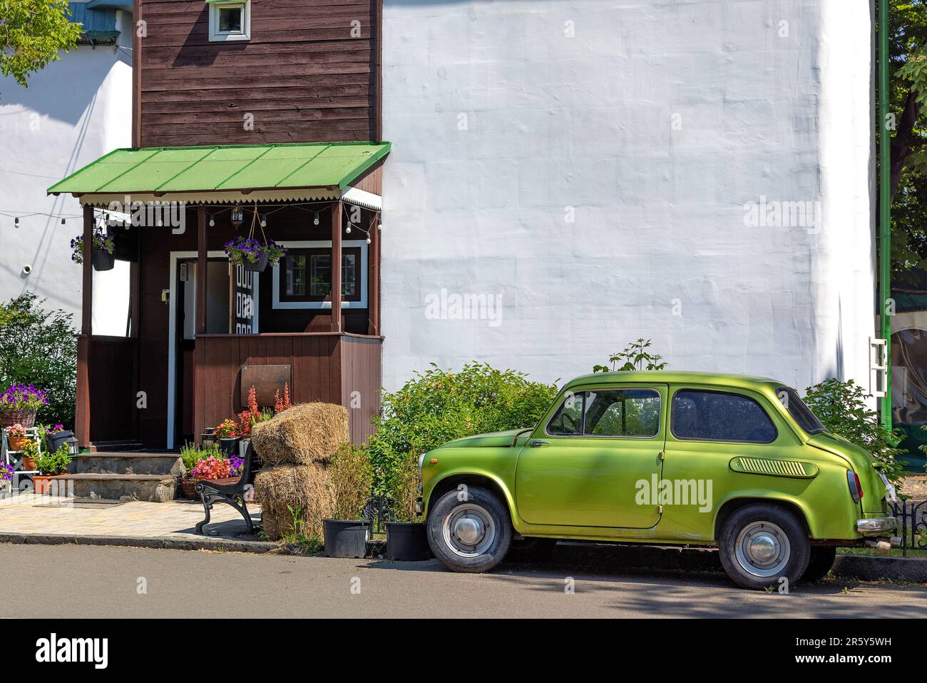 Une petite voiture verte vintage se trouve devant l'entrée d'une maison rurale avec un mur blanc et une véranda en bois par une journée ensoleillée. Copier l'espace. Banque D'Images