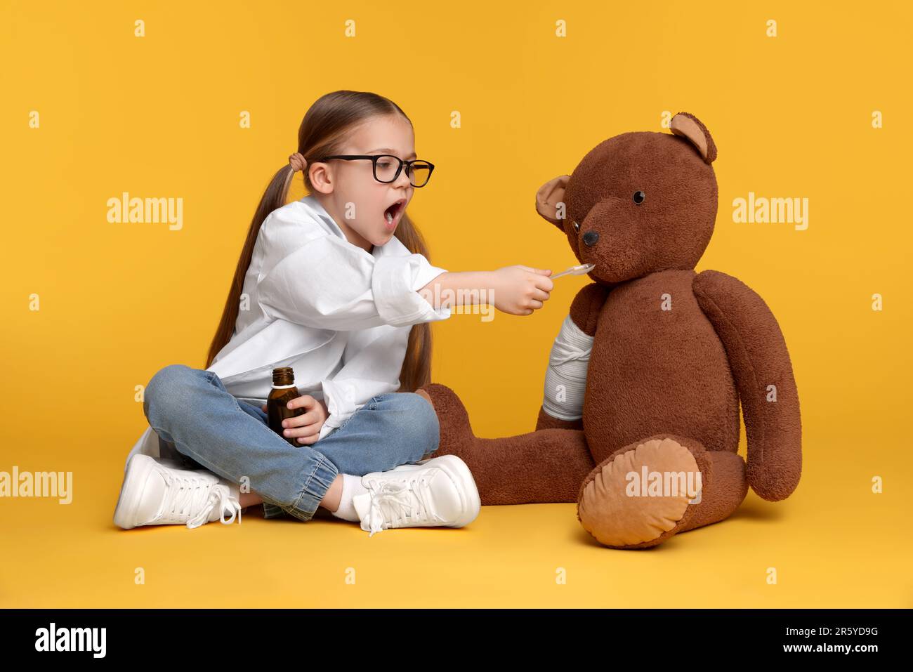 Petite fille jouant docteur avec ours jouet sur fond jaune Banque D'Images