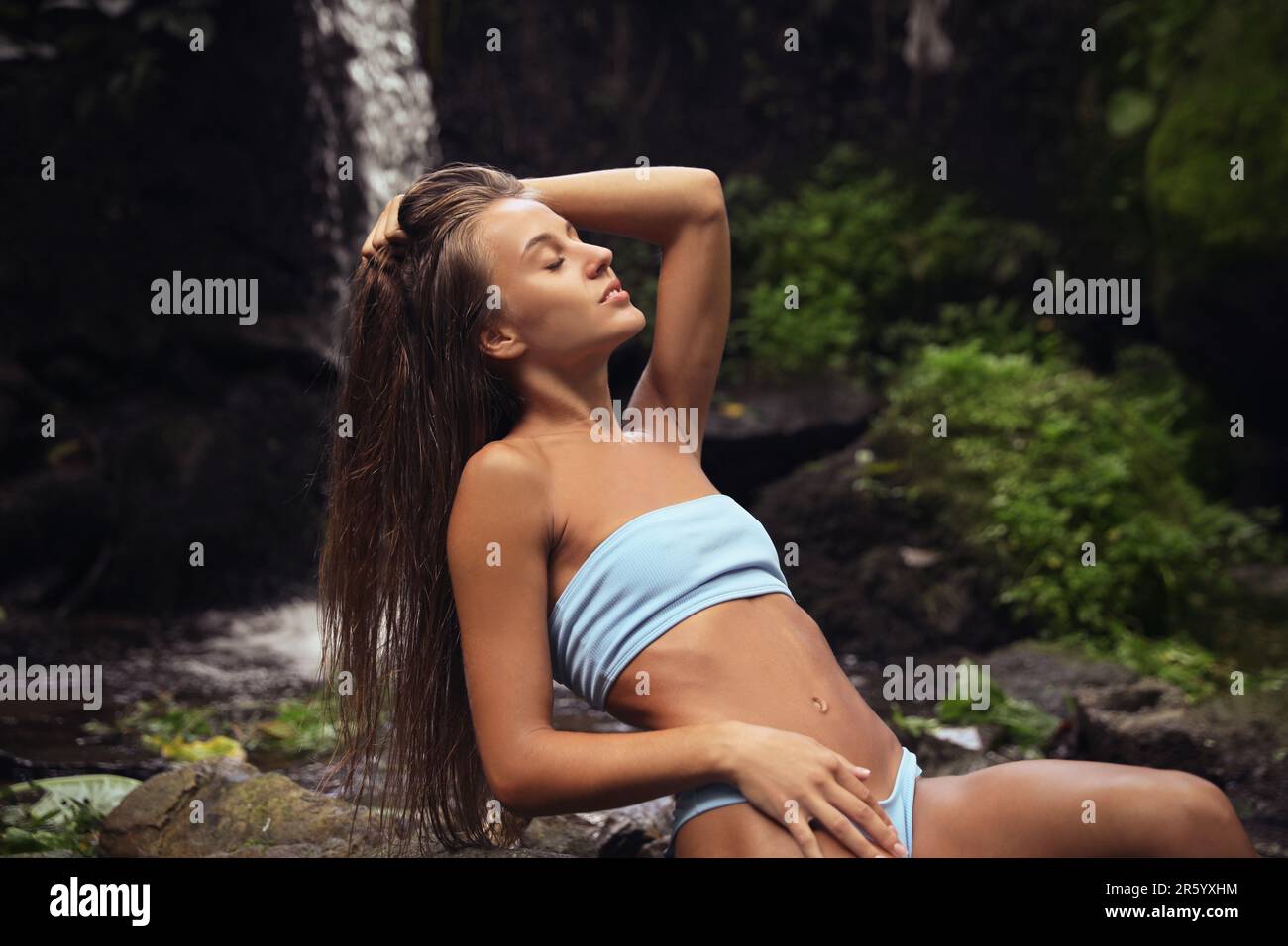 Belle jeune femme en bikini bleu clair reposant près de la cascade de montagne Banque D'Images