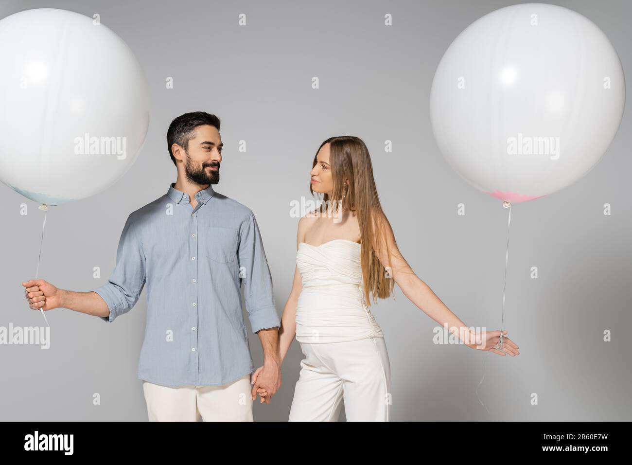 Souriant et tendance, s'attendant à ce que les parents tiennent les mains et les ballons blancs de fête tout en regardant les uns les autres pendant la fête et le sexe révèle des surprenis Banque D'Images