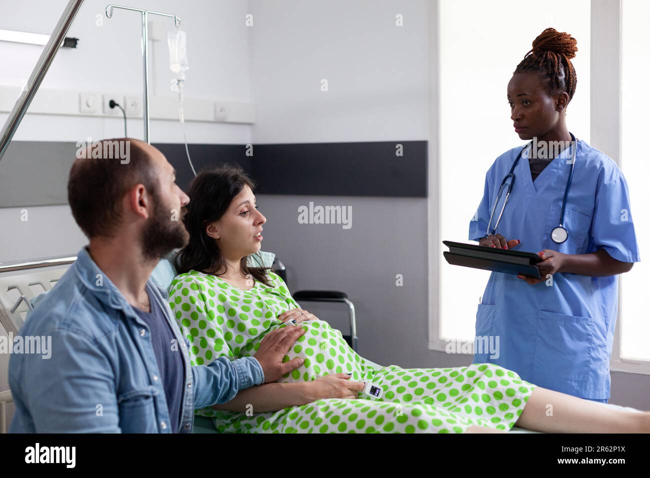 Assistant afro-américain discutant du processus d'accouchement avec la femme enceinte, préparant la mère à la chirurgie dans la salle d'hôpital. Patient avec grossesse allongé dans le lit en attente de docteur gynécologue Banque D'Images