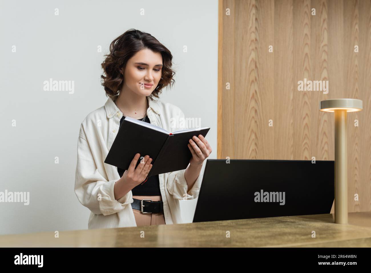 une réceptionniste positive dans des vêtements tendance et décontractés, avec des cheveux bruns ondulés et un ordinateur portable près de l'écran de l'ordinateur et de la lampe à la réception dans le hall o Banque D'Images