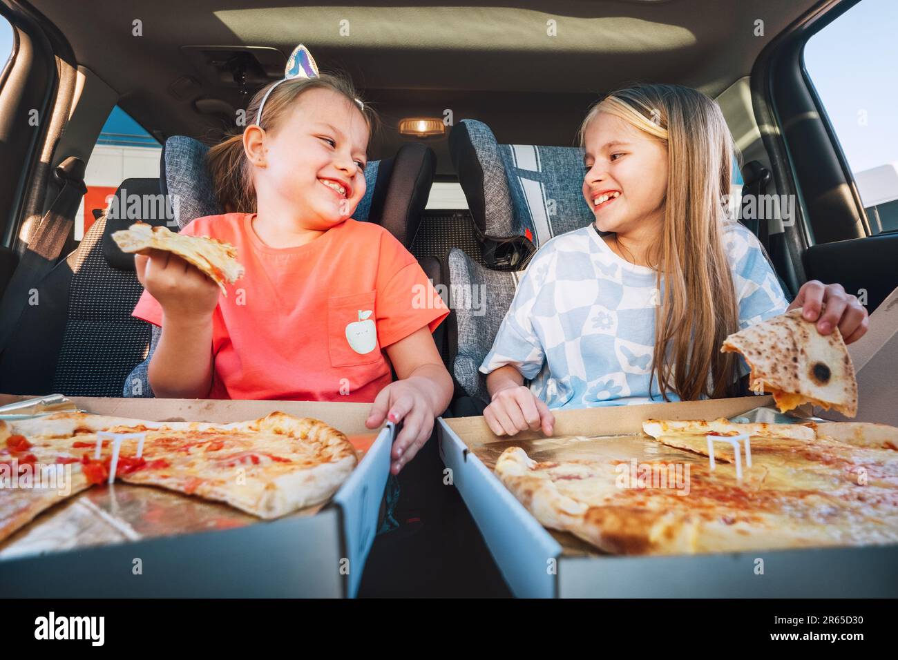 Portrait de deux sœurs souriantes et positives mangeant d'énormes pizzas italiennes juste cuites dans des sièges d'enfant sur le siège arrière de voiture. Bonne enfance, FAST-foo Banque D'Images