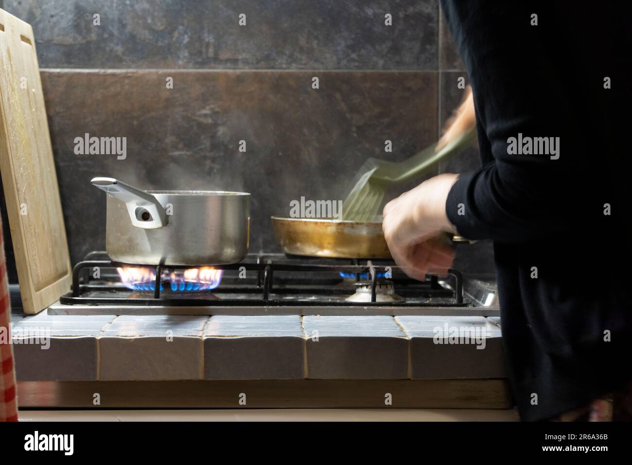 Poêle et poêle à frire sur table de cuisson à gaz utilisé pour la cuisson des repas mains de la cuisinière et des instruments de cuisson remuant cuillère juxtaposée de carrelage planche à découper en bois Banque D'Images