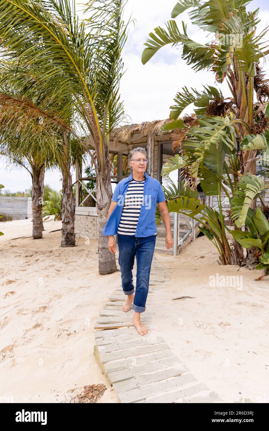 Homme âgé de race blanche marchant sur la promenade à la plage de sable contre les palmiers et le cottage Banque D'Images