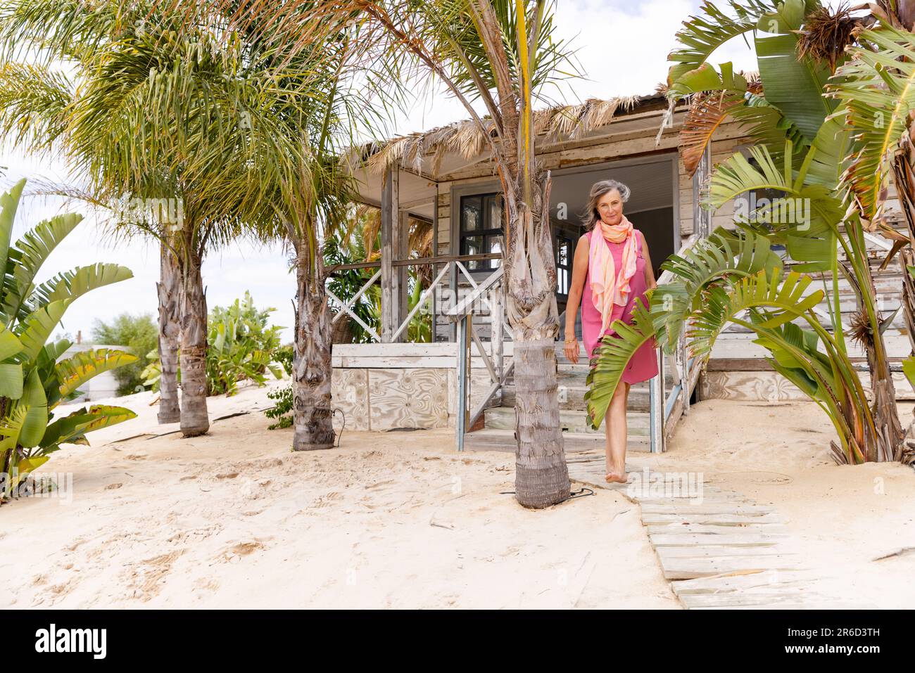 Femme caucasienne âgée marchant sur la promenade au milieu des palmiers à la plage de sable contre le cottage Banque D'Images