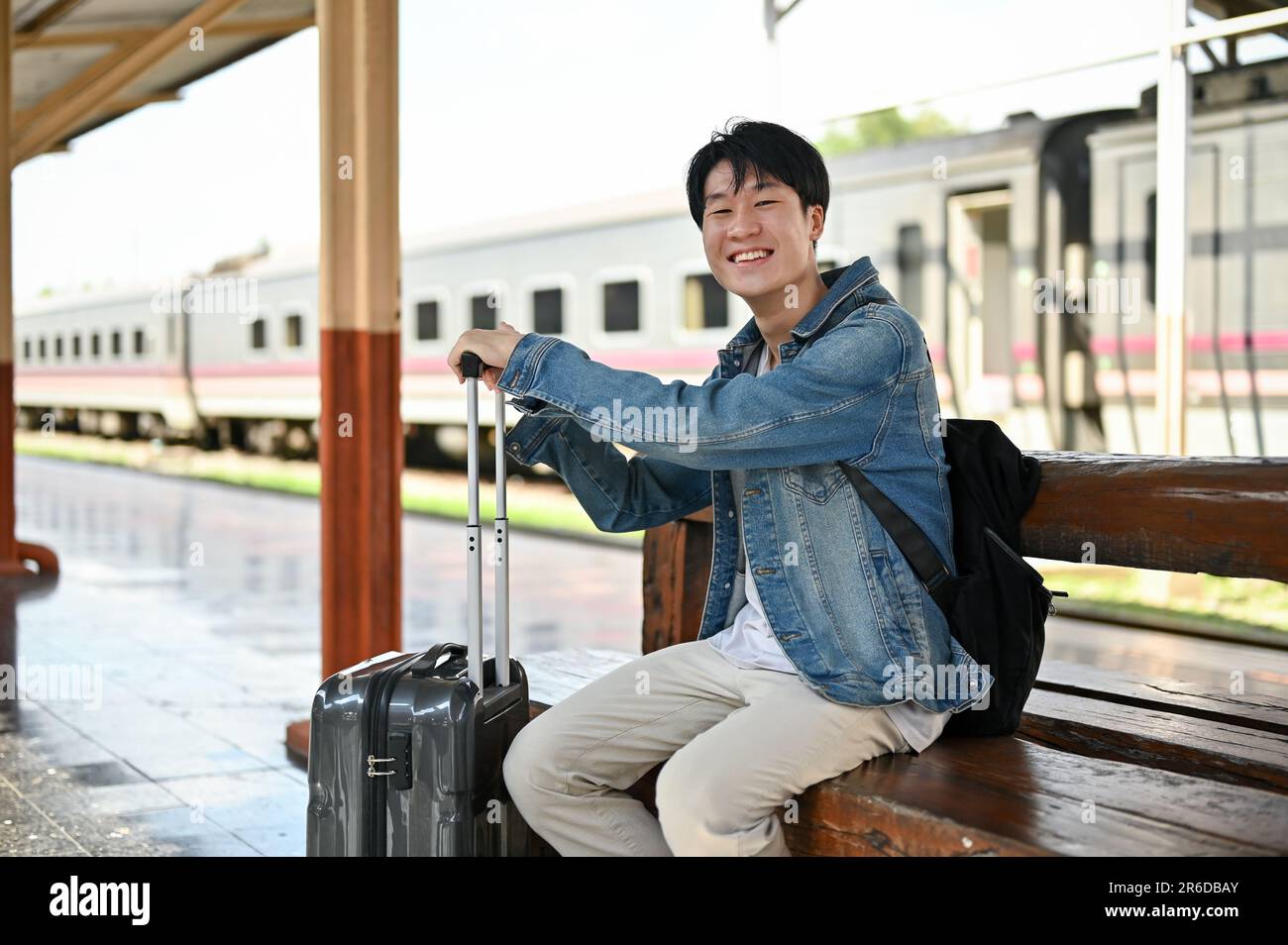 Un jeune homme asiatique souriant et heureux avec son sac à dos et sa valise est assis sur un banc en bois, attendant un train à la gare. Banque D'Images