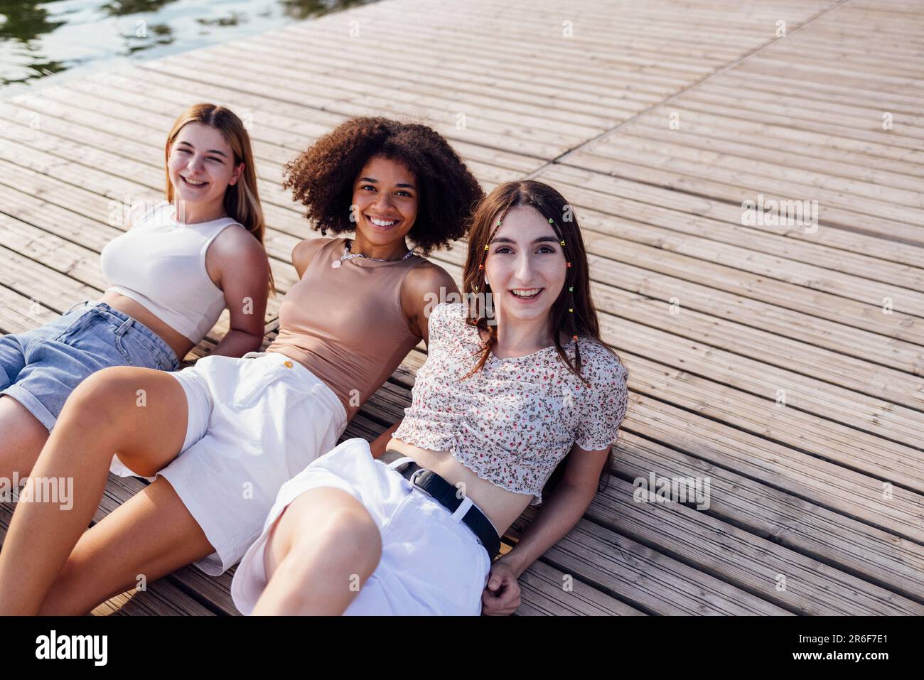 Les adolescentes souriantes et multiraciales se trouvent sur un sol en bois à l'extérieur. Rire jolies filles de différentes nationalités dans les vêtements décontractés ont grand Banque D'Images