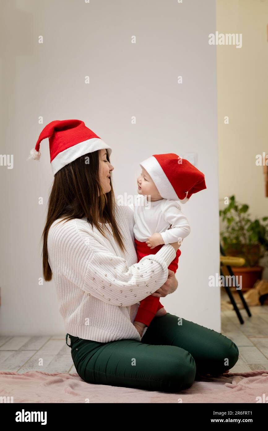 Une jeune femme tient dans les bras sa fille de 9 mois. Les deux sont dans les tenues de Noël, regardant les uns les autres et souriant heureux. Banque D'Images