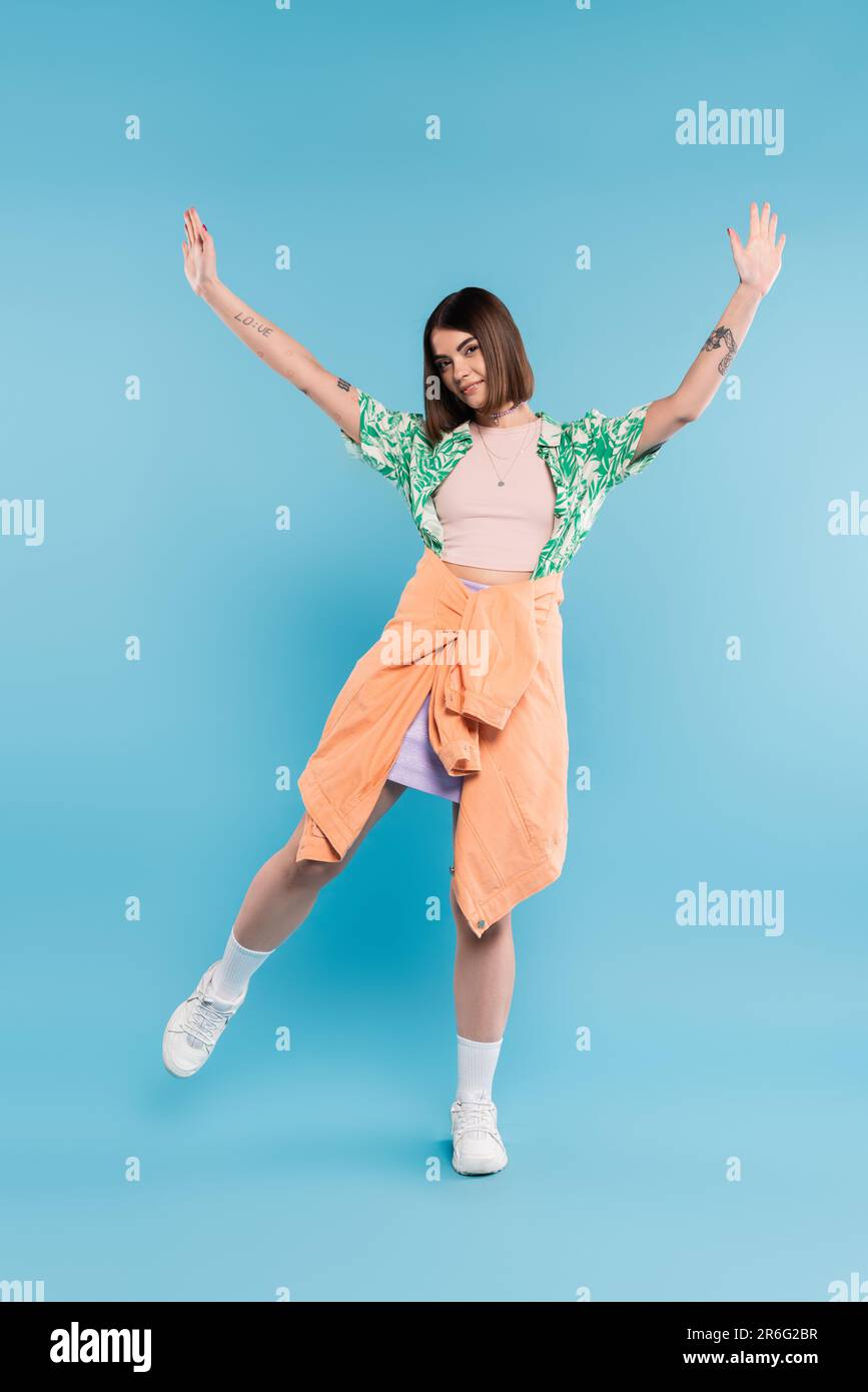 gen z mode, femme insouciante avec des cheveux courts posant dans une chemise avec imprimé palmiers, jupe et baskets blanches sur fond bleu, tatoués, pierci de nez Banque D'Images