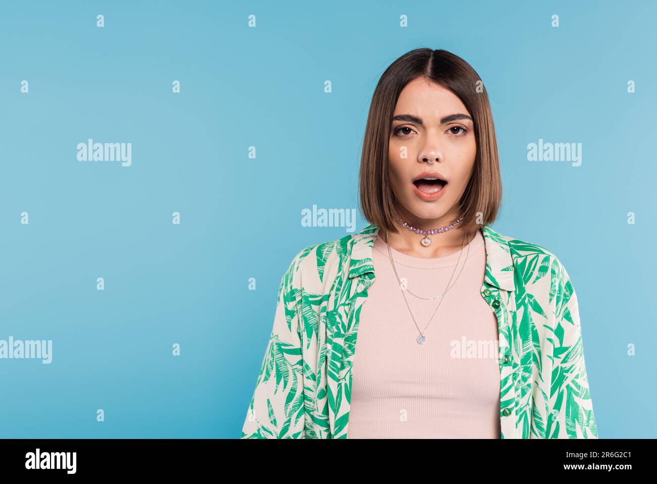femme émotif à la bouche ouverte, modèle choqué regardant la caméra sur fond bleu, chemise avec imprimé palmiers, perçage du nez, tenue décontractée, gener Banque D'Images