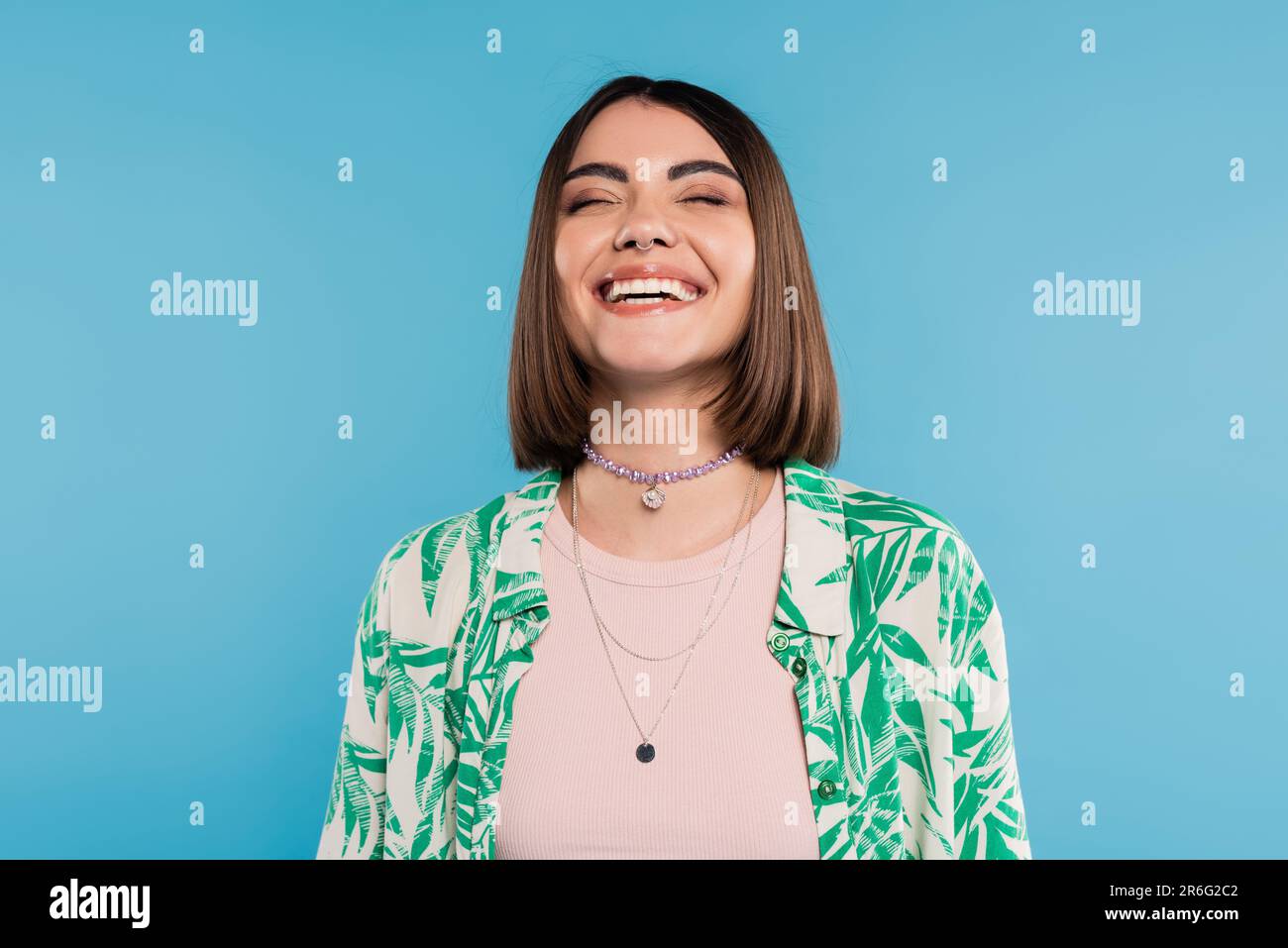 jeune femme joyeuse avec des cheveux bruns courts portant une chemise avec un imprimé palmier, souriant avec les yeux fermés sur fond bleu, tenue décontractée, gen z fash Banque D'Images
