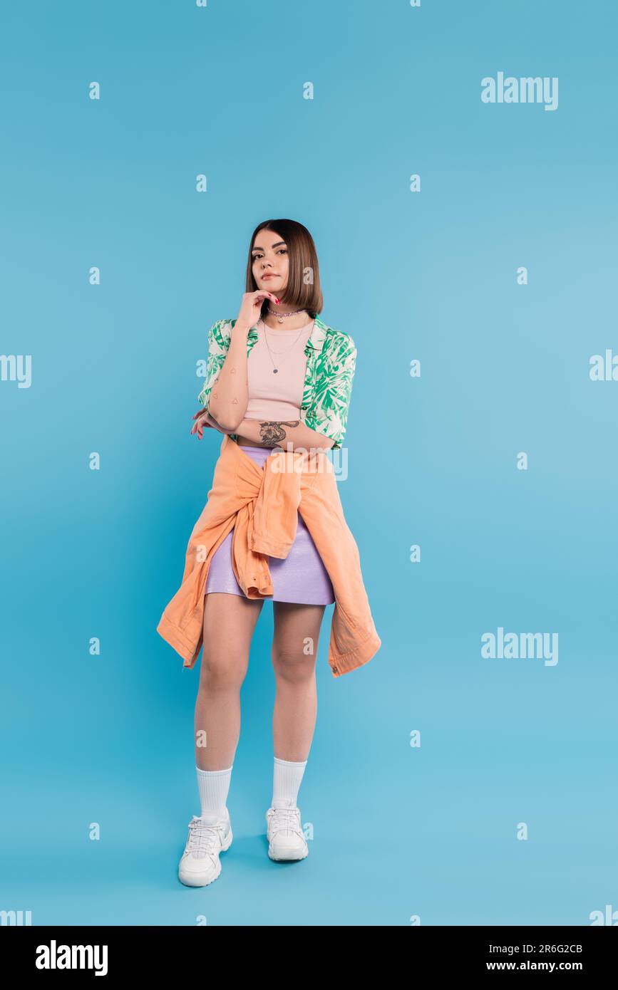 generation z, jeune femme brune aux cheveux courts posant dans une chemise avec imprimé palmiers, jupe et baskets blanches sur fond bleu, tatouées, nez p Banque D'Images