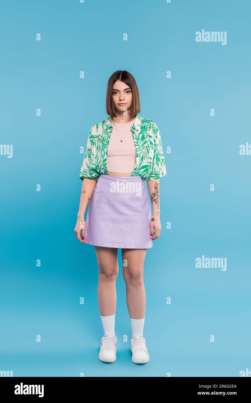 gen z mode, jeune femme brune aux cheveux courts posant dans une chemise avec imprimé palmiers, jupe et baskets blanches sur fond bleu, insouciante, tatto Banque D'Images