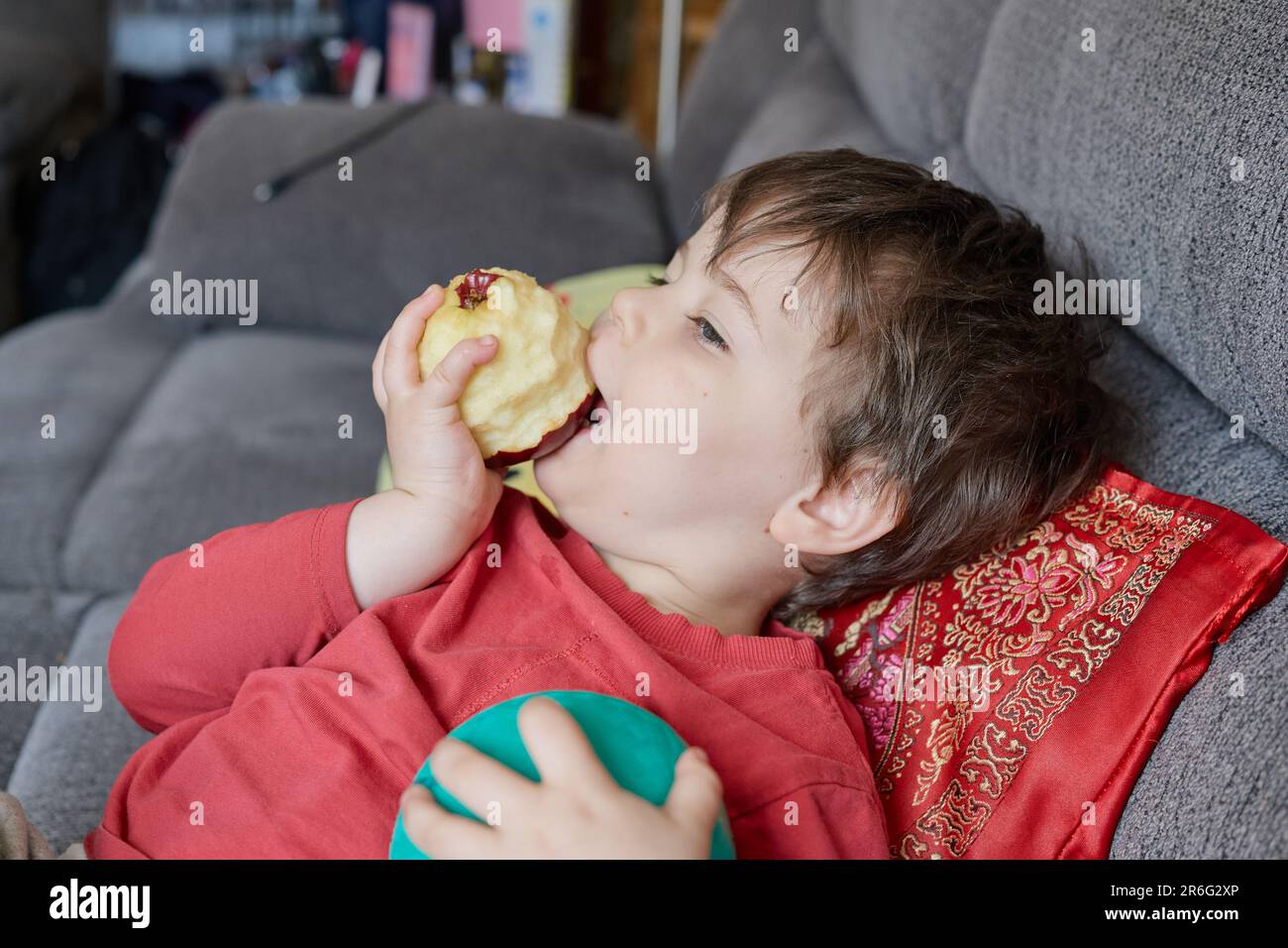 jeune garçon expressif mangeant une pomme et se prélassant sur le canapé à la maison Banque D'Images