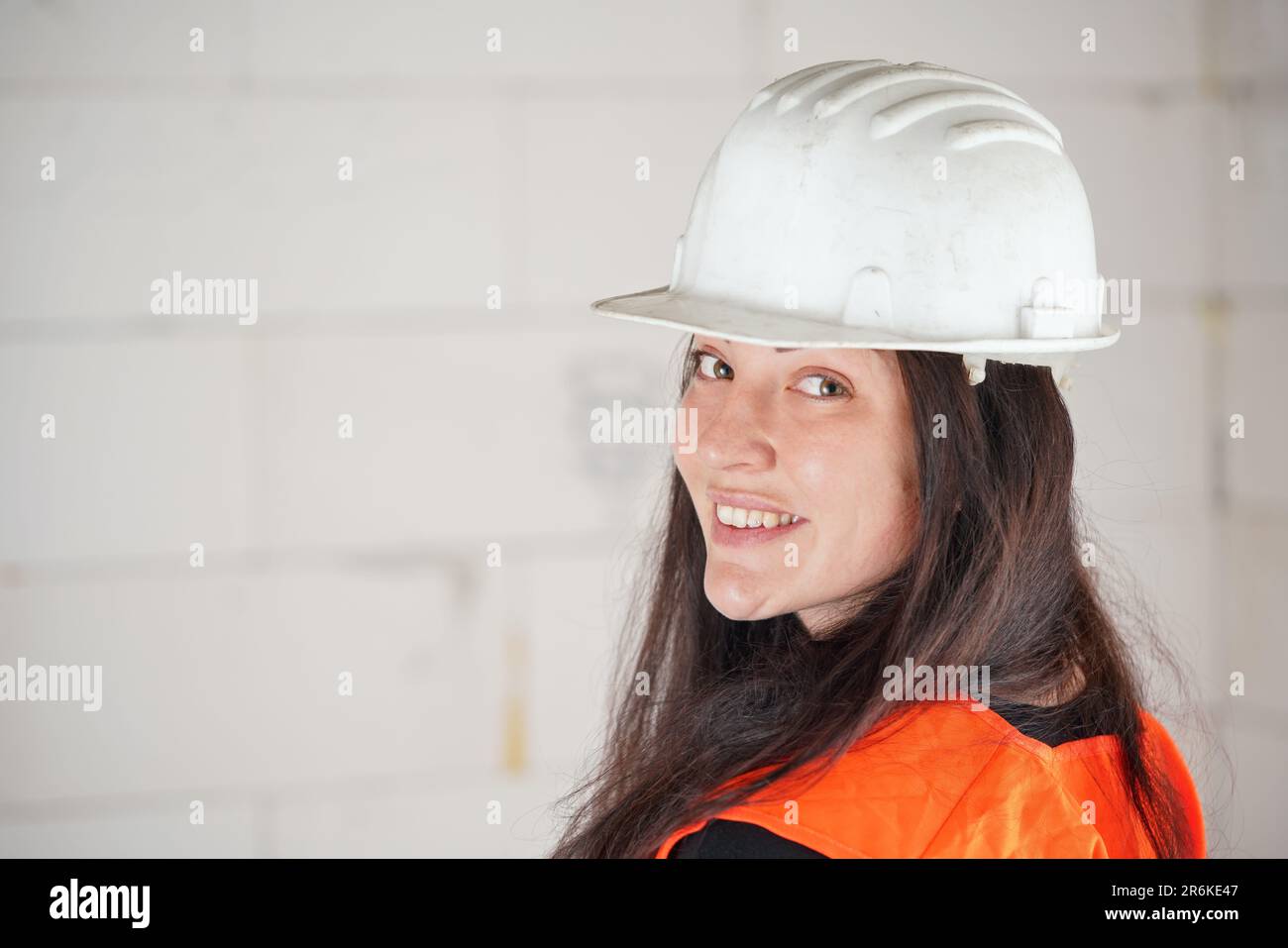 Jeune femme en casque blanc et gilet orange haute visibilité, cheveux longs foncés, regardant par-dessus son épaule dans l'appareil photo, souriant. Construction floue si Banque D'Images