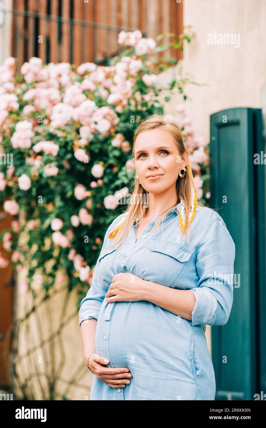 Portrait extérieur de femme enceinte élégante posant dans un jardin de roses, portant une robe en denim Banque D'Images