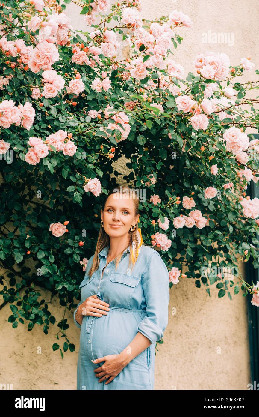 Portrait extérieur de femme enceinte élégante posant dans un jardin de roses, portant une robe en denim Banque D'Images