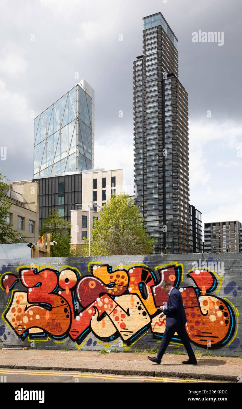 Londres, Royaume-Uni - 17 mai 2023: Un homme d'affaires en costume marche à côté d'un mur couvert de graffiti à Shoreditch, engrosé dans son téléphone, avec les grands bâtiments de Banque D'Images