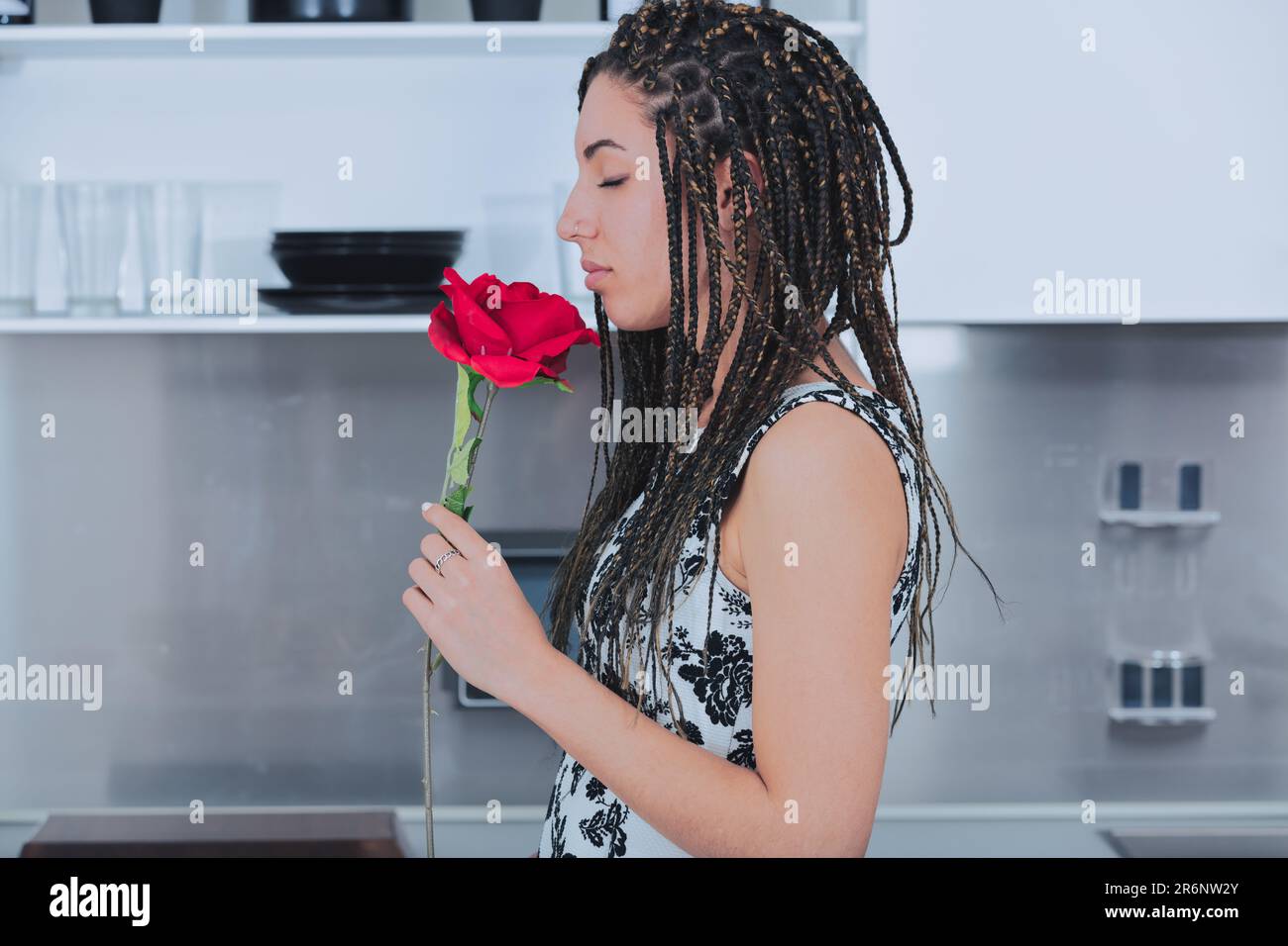 Dans sa cuisine, une fille avec boîte de cheveux tressés passionément sniffs une rose. Cette jeune femme porte une robe florale et sports divers bijoux perceur. Elle Banque D'Images