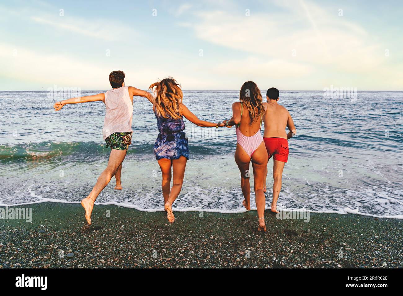 Quatre jeunes amis, deux couples caucasiens, courent vers la mer calme pour une baignade rafraîchissante. La caméra les capture de la plage, avec leur dos t Banque D'Images
