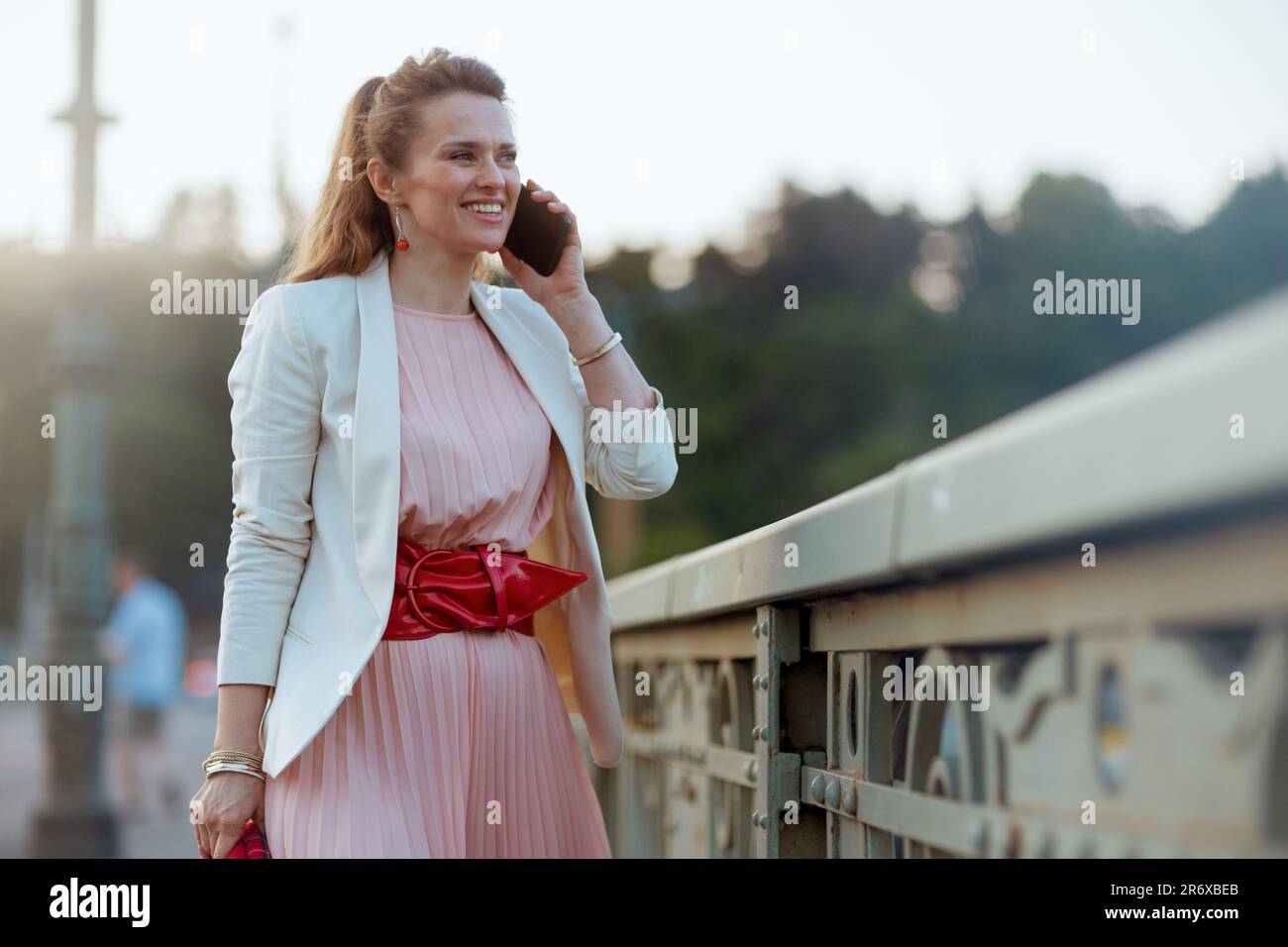 souriante élégante femme de 40 ans en robe rose et blouson blanc dans la ville parlant sur un smartphone sur le pont. Banque D'Images