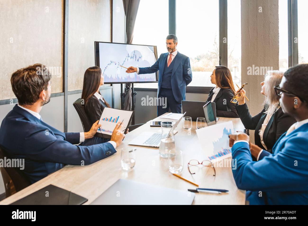Un dirigeant de sexe masculin pointe vers l'écran et explique les tableaux financiers à ses collègues lors d'une réunion d'affaires dans une salle de réunion ensoleillée Banque D'Images
