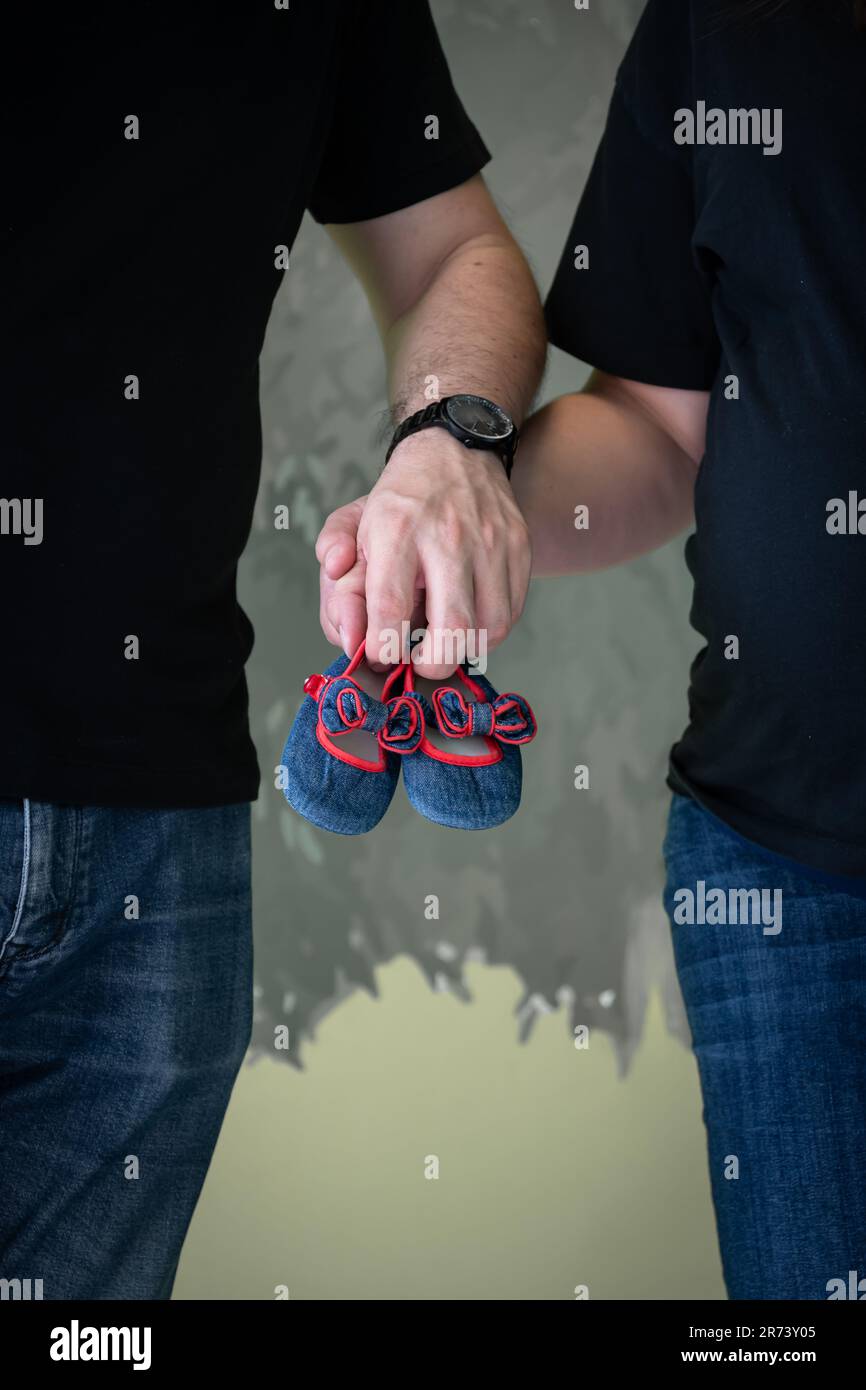Femme enceinte et son mari, vêtus de jeans et d'un t-shirt noir, tiennent les chaussures de bébé dans leurs mains. Banque D'Images