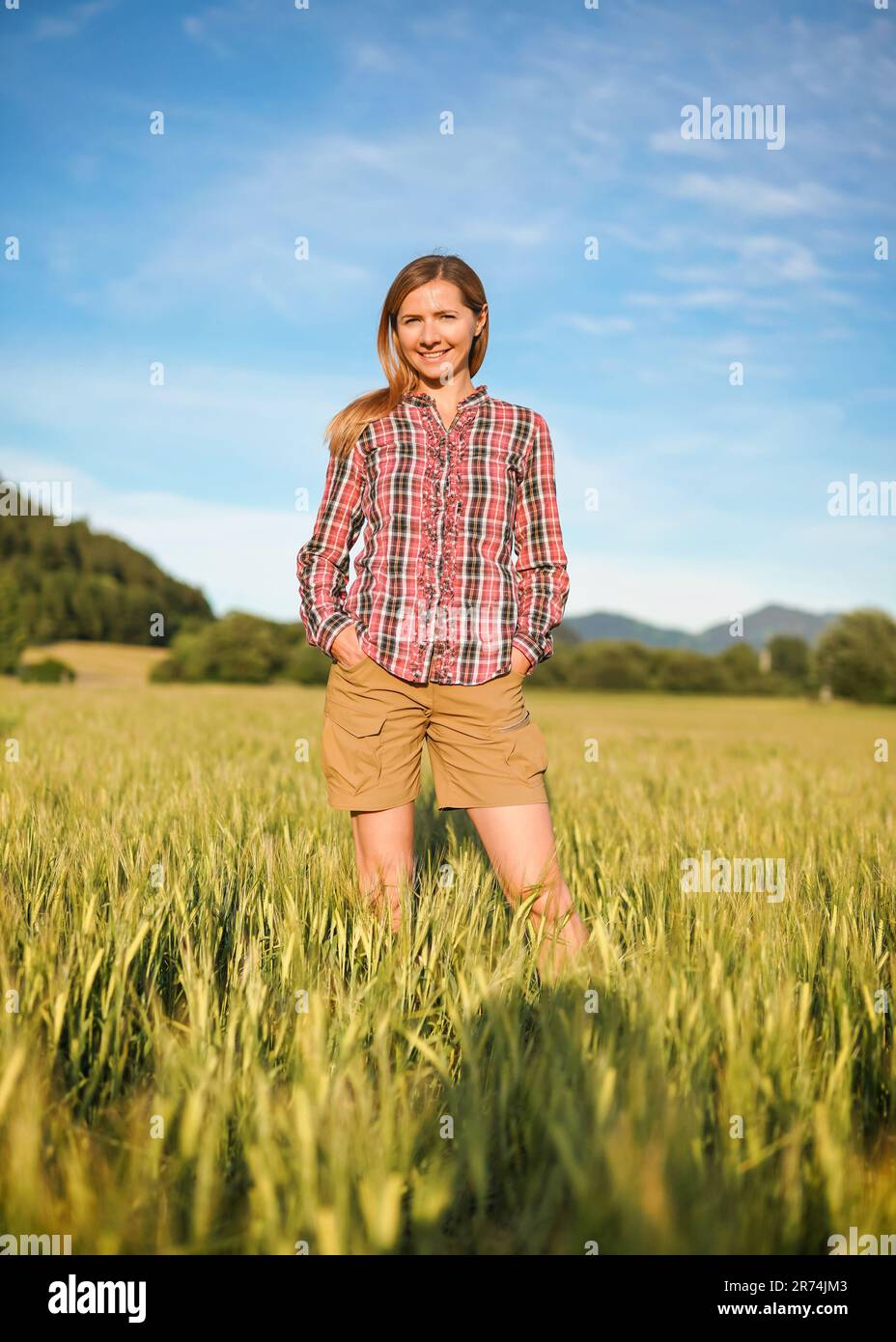 Jeune femme en chemise à carreaux Vichy et short debout dans un champ de blé vert non mûr, sourire, ciel bleu et paysage flou avec petit dos de forêt Banque D'Images