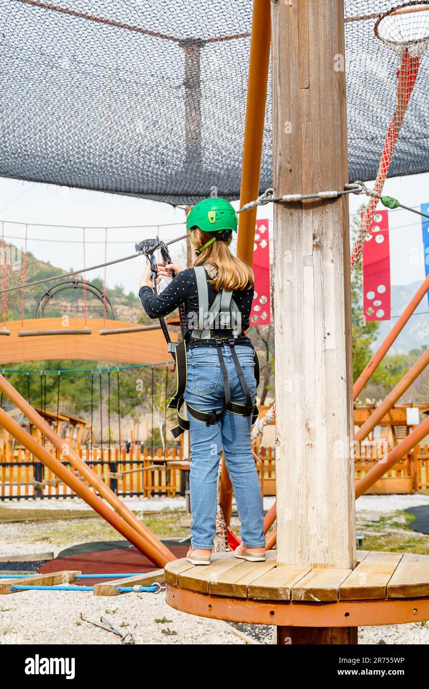 Adolescente dans l'équipement de harnais d'escalade, casque de sécurité vert de sport. Parc d'attractions de corde. Fixation fixant le mousqueton à la corde de sécurité. Hangin Banque D'Images