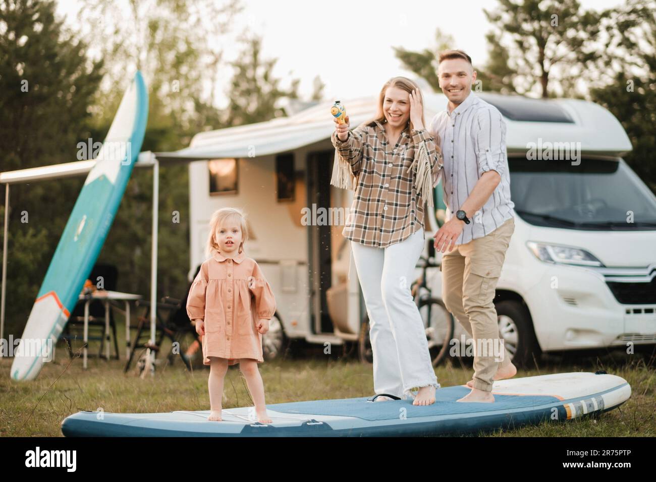 la famille se repose à côté de sa maison mobile. Papa, maman et fille jouent sur des planches de sup avec des pistolets d'eau près de la motorhome. Banque D'Images