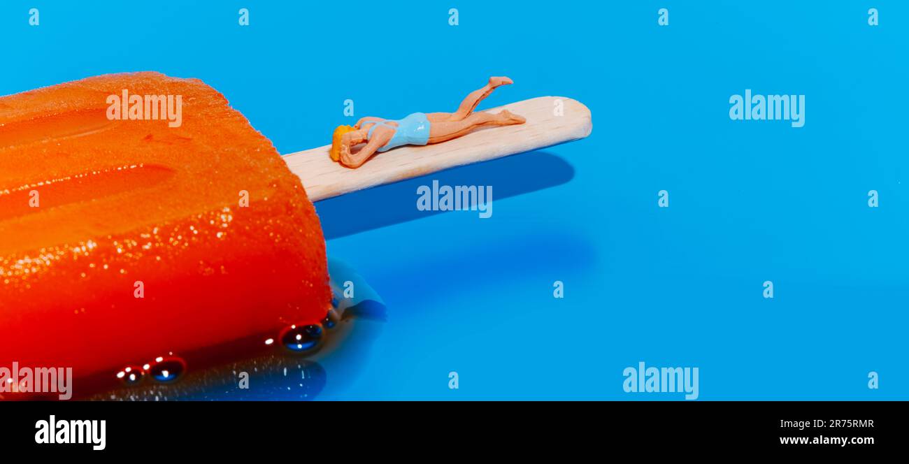 gros plan d'une femme miniature, vêtue d'un maillot de bain bleu, allongé sur le pôle d'un pop-sicle orange en fusion sur fond bleu, dans une forme panoramique Banque D'Images