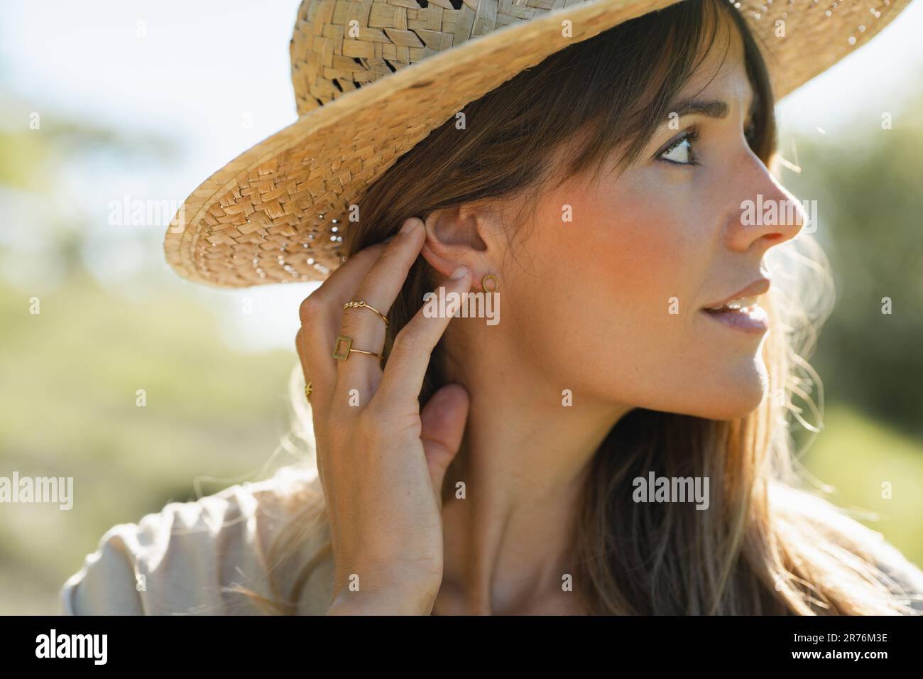Portrait d'une jeune femme souriante en chapeau de paille touchant l'arrière de la tête avec les doigts et regardant loin contre un arrière-plan flou en plein soleil Banque D'Images