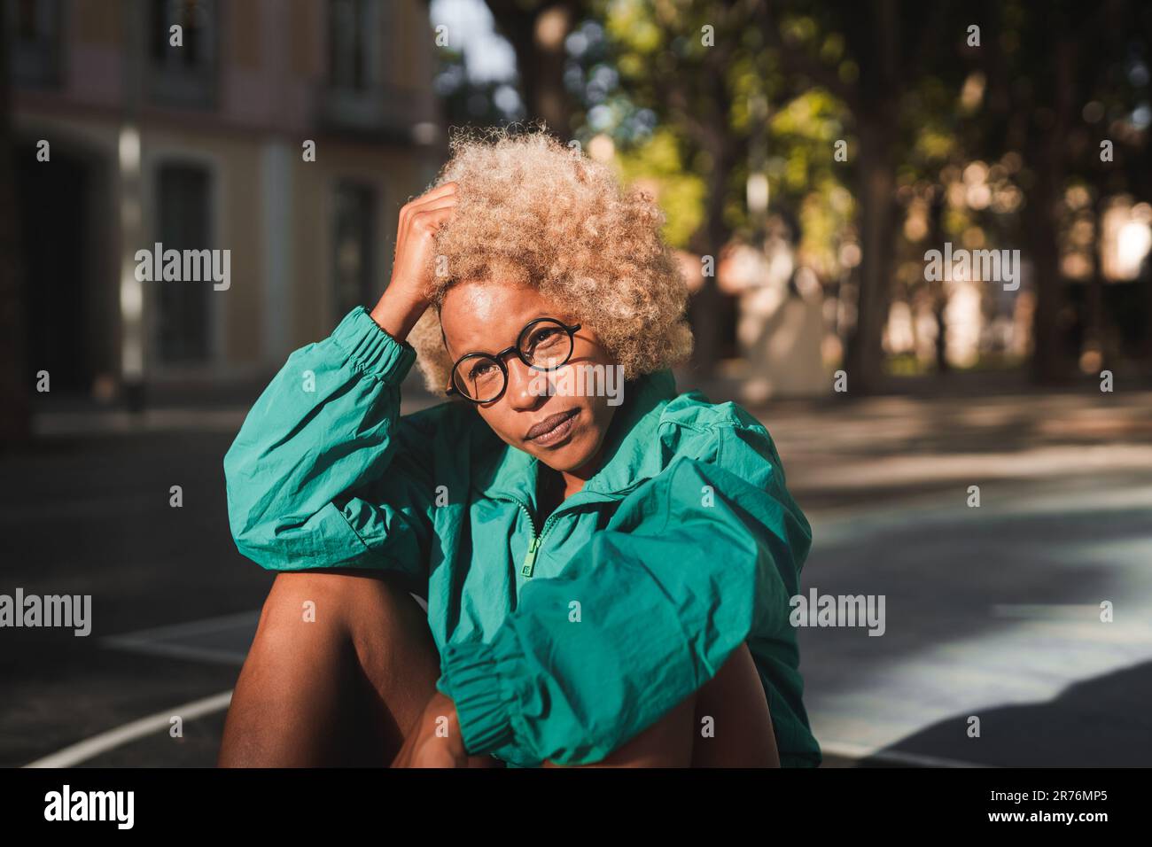 Bonne jeune athlète afro-américaine en vêtements de sport avec lunettes assises sur le terrain de basket-ball au coucher du soleil Banque D'Images