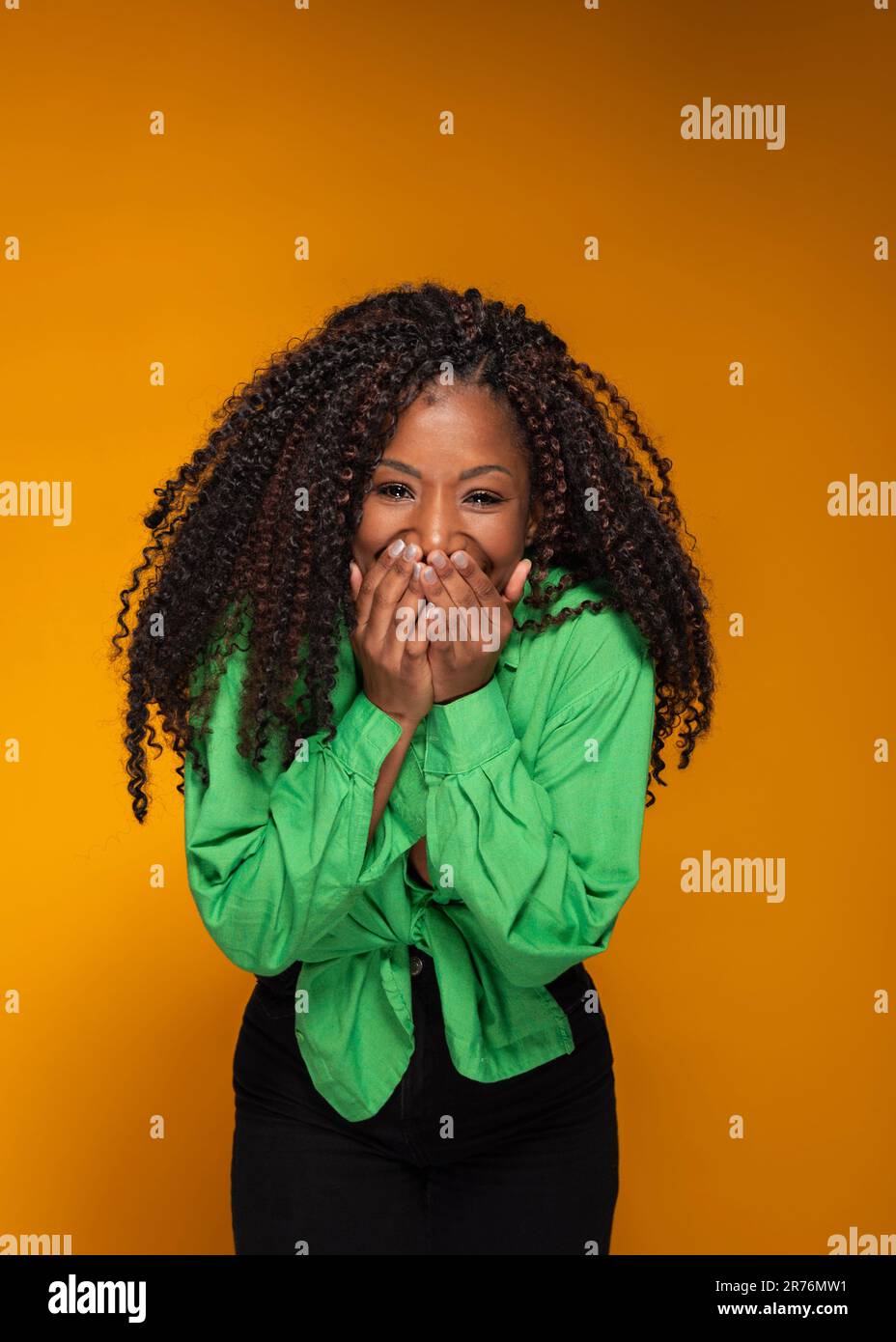 Portrait d'une femme noire ravie dans des vêtements élégants souriant et couvrant le visage tout en regardant l'appareil photo sur fond orange Banque D'Images