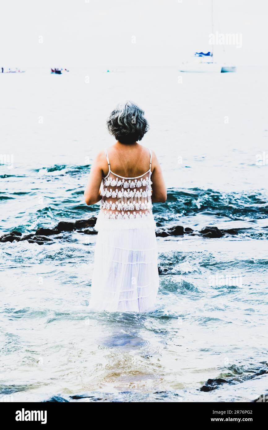 Une femme mûre aux cheveux gris foncé est debout sur une plage rocheuse portant une robe en crochet blanc Banque D'Images