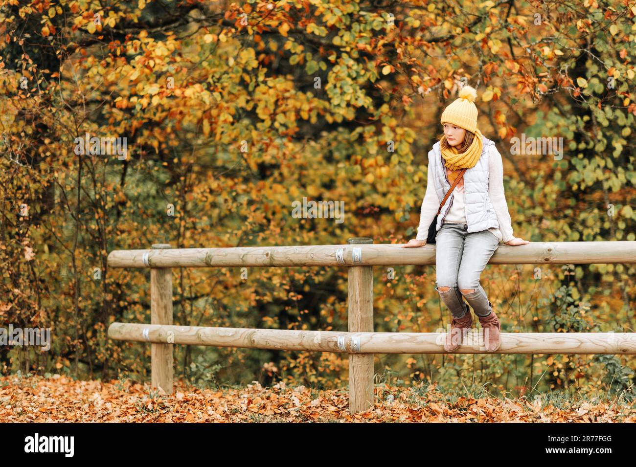 Portrait d'automne d'une adorable jeune fille de 10-11 ans, portant une veste en duvet grise, un chapeau et une écharpe jaunes, assis sur la clôture Banque D'Images