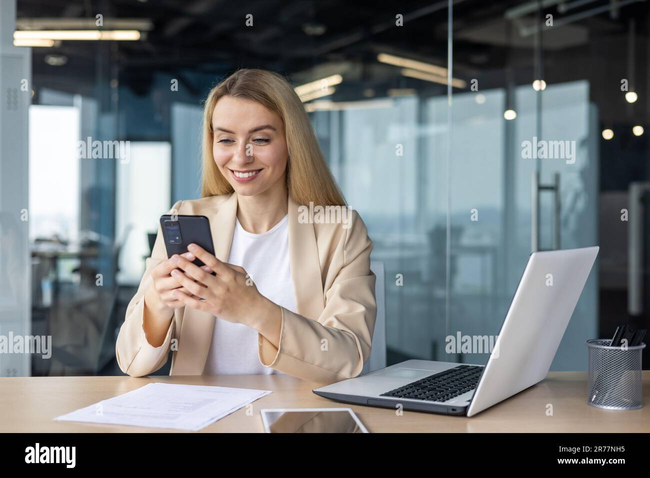 Belle femme d'affaires réussie et heureuse travaillant à l'intérieur du bureau sur le lieu de travail, blonde tenant le téléphone en utilisant l'application en ligne, souriant assis à l'onglet Banque D'Images