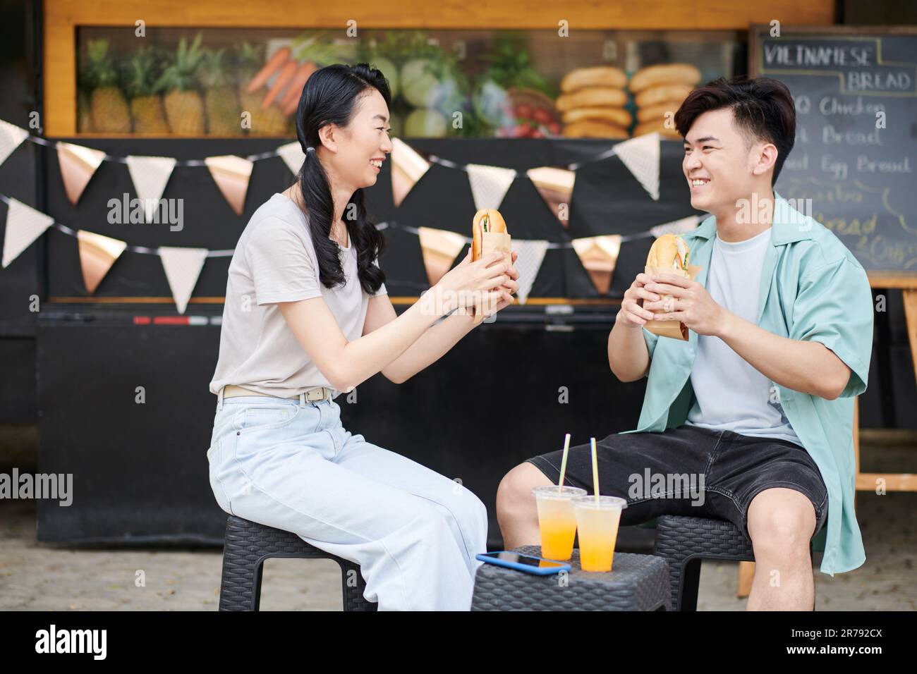 Jeunes asiatiques joyeux dattes en vêtements décontractés ayant de savoureux hot dogs à l'extérieur tout en étant assis contre un camion de restauration rapide de la rue pendant la pause et le chat Banque D'Images