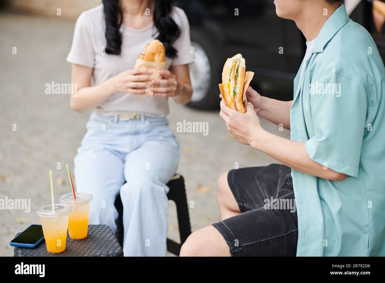 Petit cliché de jeune homme en tenue décontractée ayant appétissant hot dog tout en étant assis devant sa petite table et sa petite petite petite table avec deux jus d'orange Banque D'Images