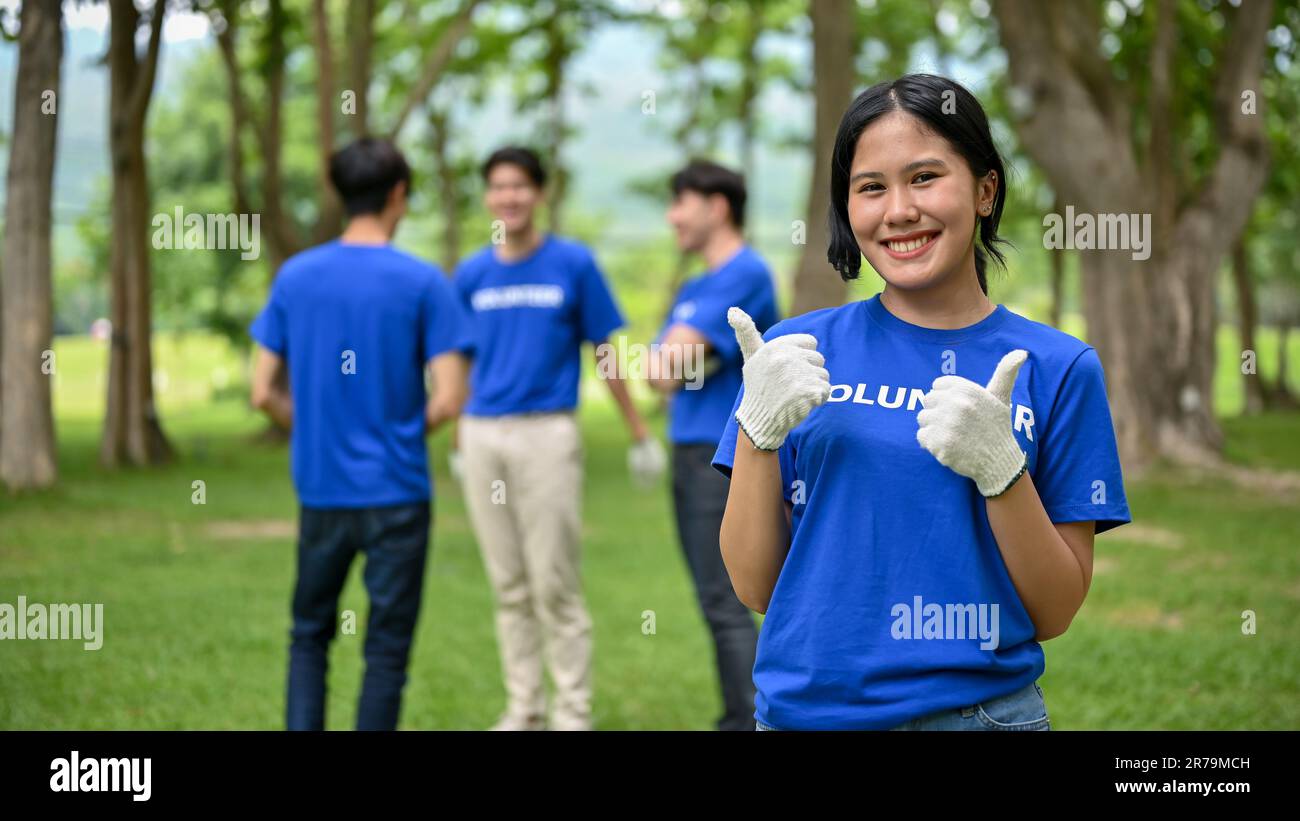 Une jeune femme asiatique, belle et heureuse, se tient dans un parc public et montre ses pouces, participant au travail bénévole avec ses amis t Banque D'Images