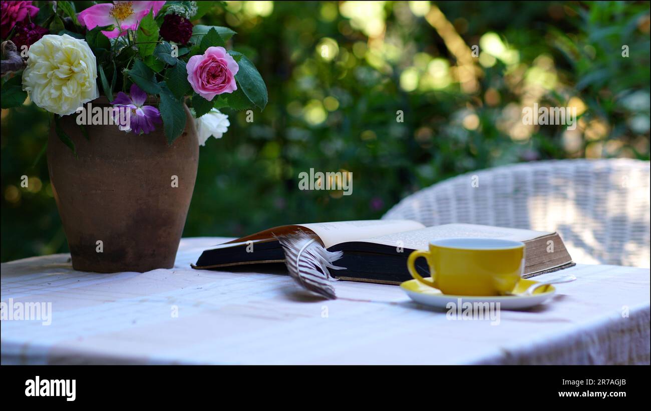 Un coin salon confortable dans le jardin. Un bouquet de roses sur une table, un livre ouvert avec une plume, une tasse jaune. Une chaise en osier. Banque D'Images