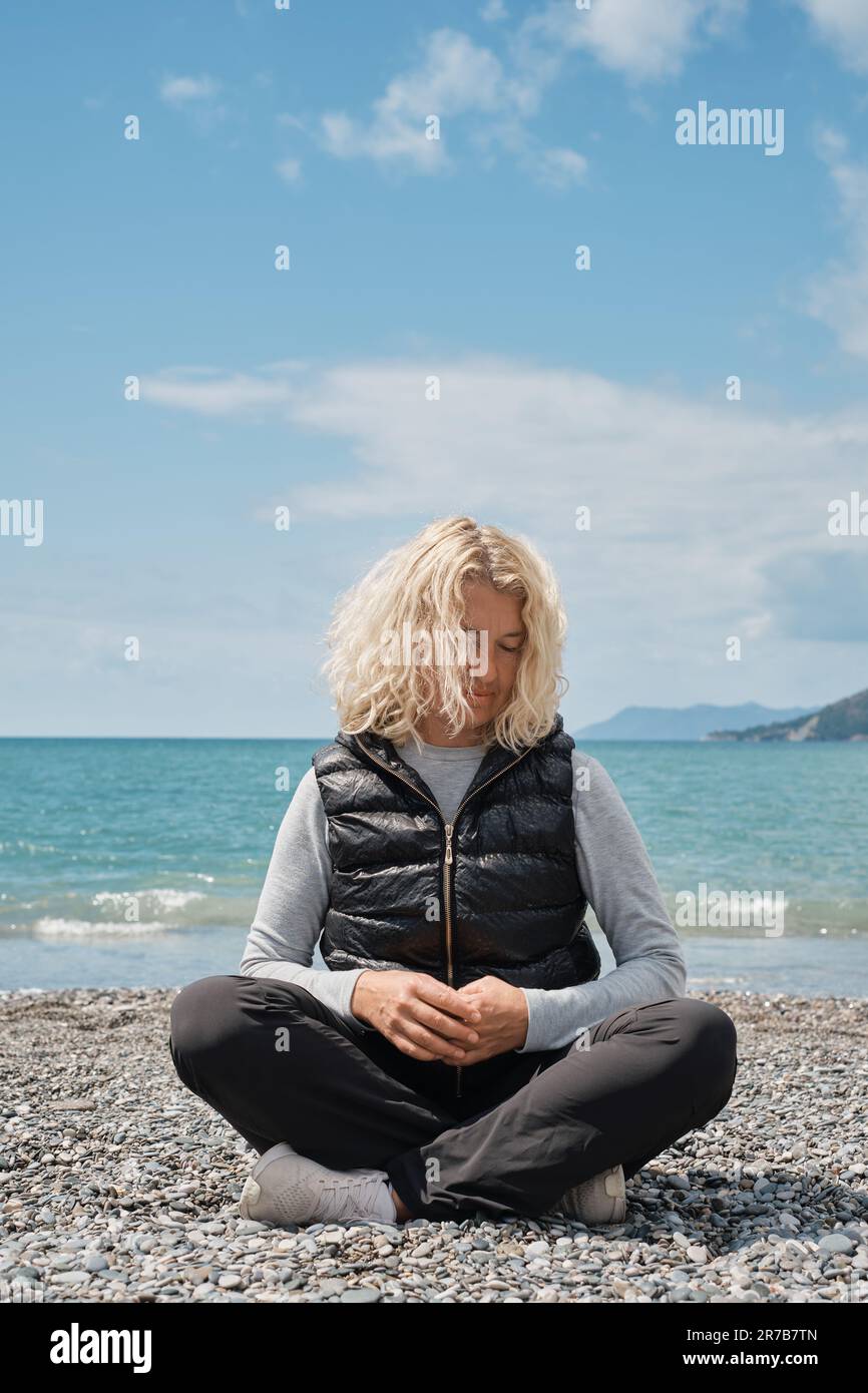 Adulte touriste européenne femme blonde assise seule sur une plage de galets avec bleu océan et le ciel en arrière-plan, tir vertical. Les gens et l'été Holida Banque D'Images