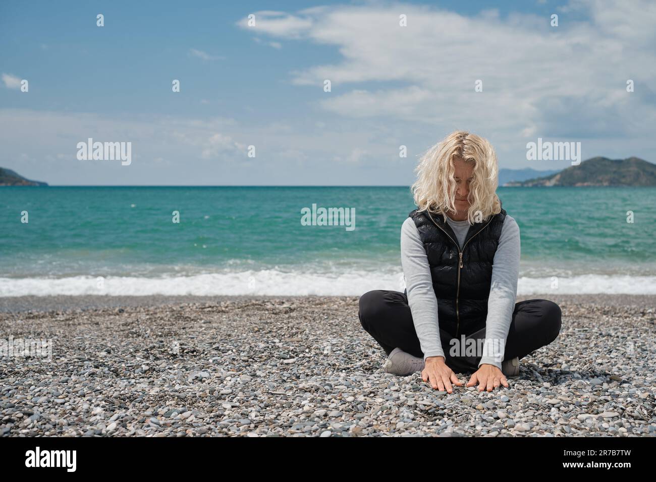 une femme blonde européenne est assise sur une plage de galets un jour de printemps touche les pierres de la plage avec ses mains. Portrait extérieur d'une belle adu curly Banque D'Images