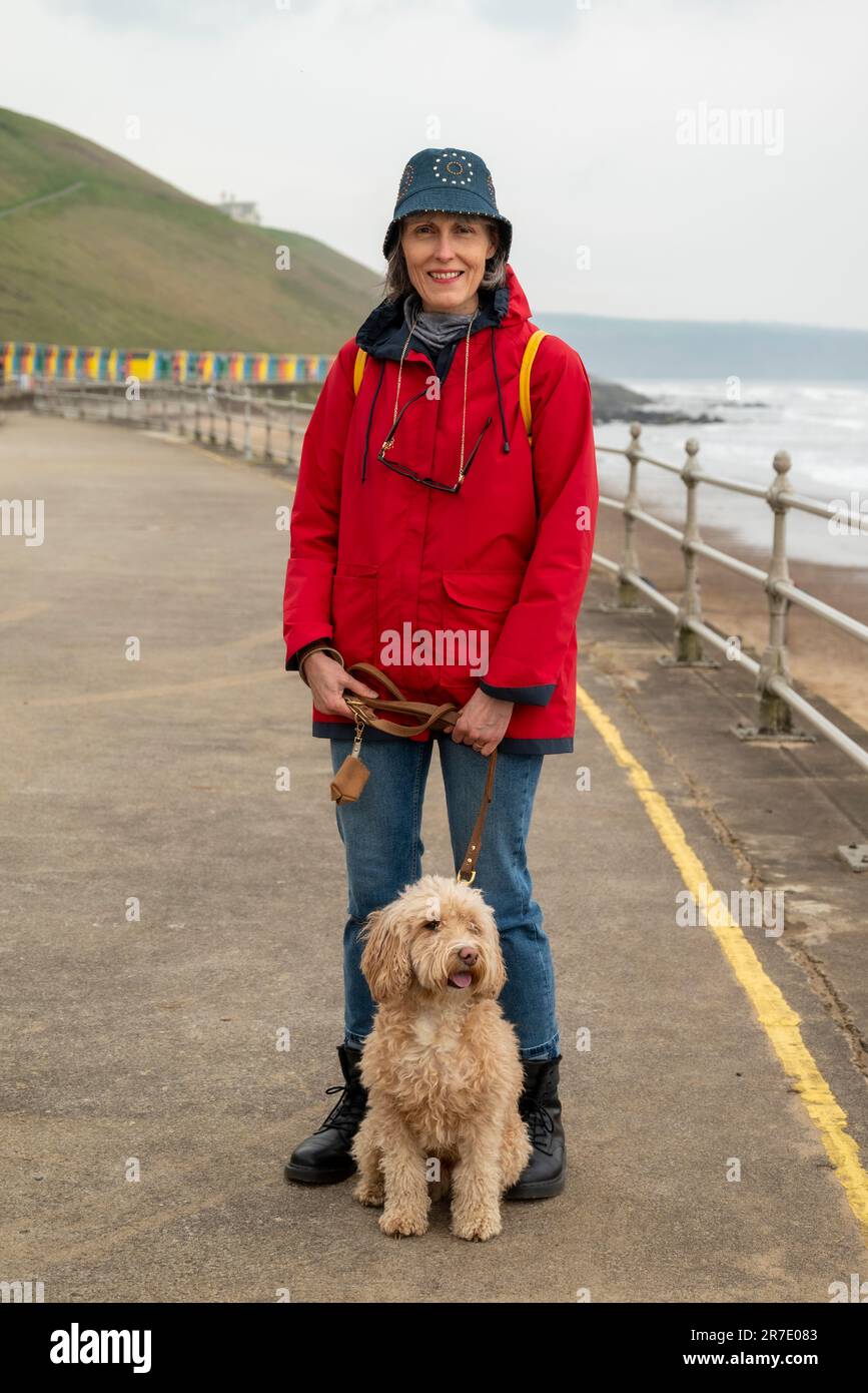 Femme caucasienne mûre souriante vêtue d'un manteau rouge imperméable et d'un chapeau de seau bleu debout avec un cocapoo blond sur le front de mer de Whitby. Banque D'Images