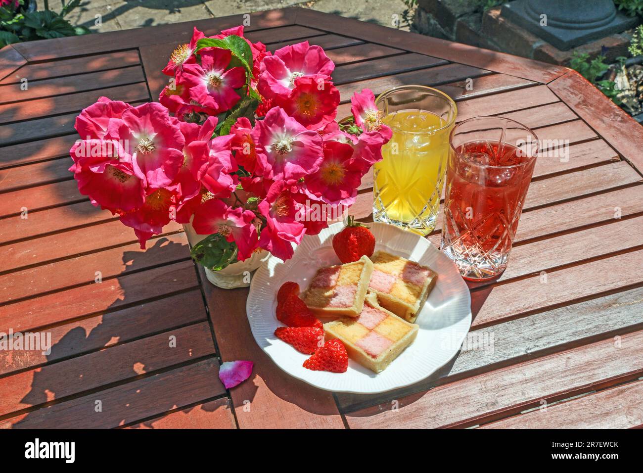 Une assiette de tranches de gâteau de battenberg sur une table de jardin en lamelles avec dronks doux et aposy de roses en rameau; Banque D'Images