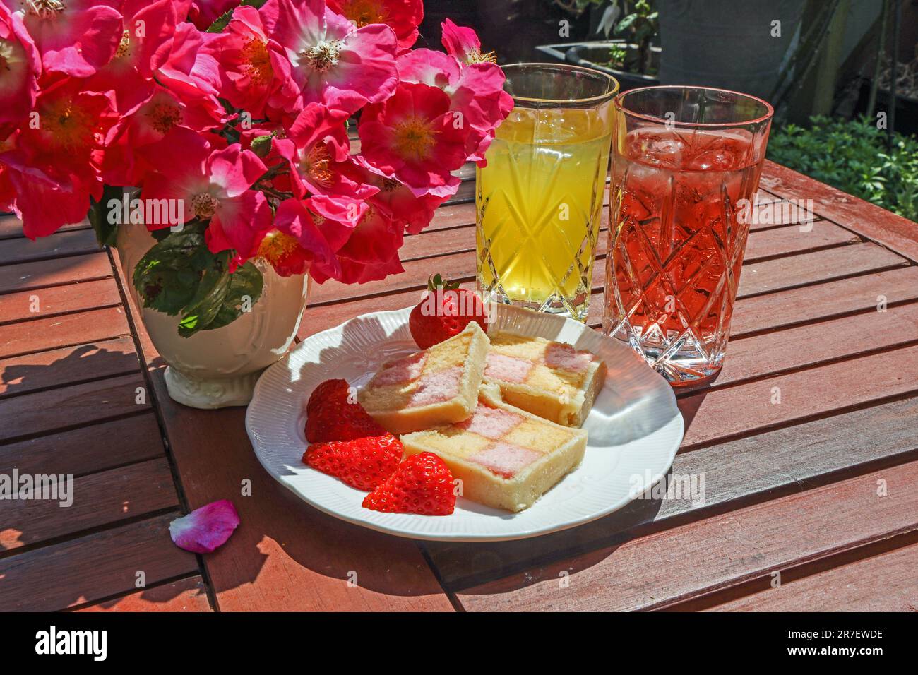 Une assiette de tranches de gâteau de battenberg sur une table de jardin en lamelles avec dronks doux et aposy de roses en rameau; Banque D'Images