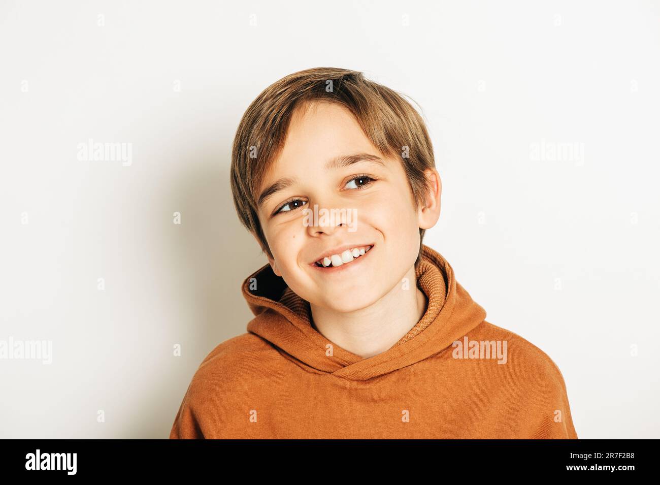 Photo studio d'un beau garçon de 10 ans aux cheveux blonds, portant un sweat à capuche marron, posant sur fond blanc, regardant vers le haut Banque D'Images