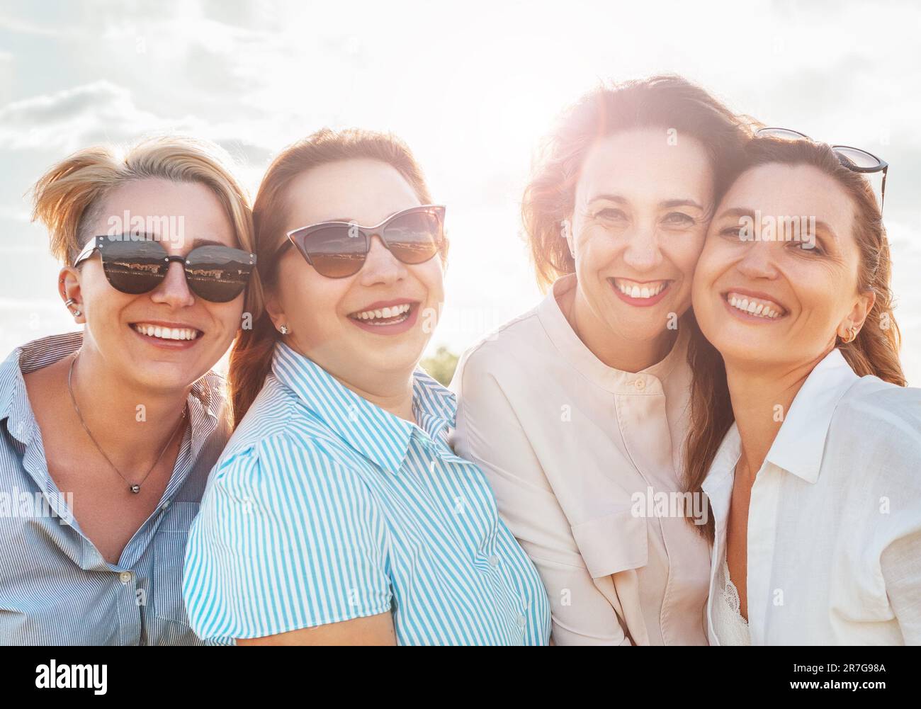 Portraits en demi-longueur de quatre femmes souriantes en lunettes de soleil embrassant et regardant l'appareil photo pendant la marche en plein air avec un soleil éclatant. ven femme Banque D'Images